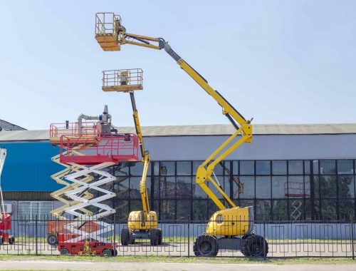 Several different wheeled scissor lifts and wheeled articulated lifts with telescoping boom and basket on an asphalt ground against the sky and an industrial building