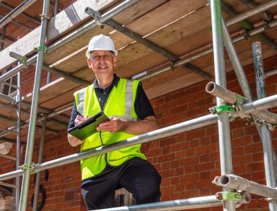 Male builder foreman, construction worker, contractor or architect on building site writing on black clipboard laughing, smiling and happy